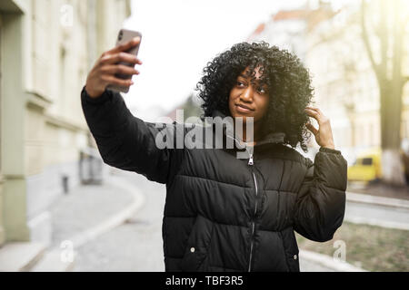 Cheerful young African woman wearing coat la marche à l'extérieur, en prenant un selfies Banque D'Images