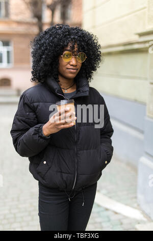 Cheerful young African woman wearing coat la marche à l'extérieur, tenant la tasse de café à emporter Banque D'Images