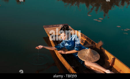 La fille est allongée sur un petit bateau en bois sur la surface du lac, s'emparer de l'eau du lac avec ses mains, l'eau s'égouttant de ses doigts, la hat Banque D'Images