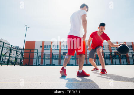 Deux Caucasian friends wearing red matériel sport basket jouer à l'extérieur sur une journée ensoleillée alors qu'ils dribble et pousser l'autre. Banque D'Images