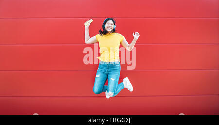 Happy Asian girl jumping tout en écoutant la musique en plein air - Crazy femme chinoise s'amuser danser une chanson contre l'arrière-plan rouge Banque D'Images