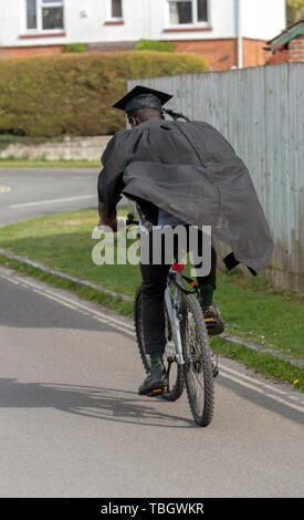 Andover, Hampshire, England, UK. Mai 2019. Une vue arrière d'un étudiant à l'université wearing cap and gown la bicyclette. Banque D'Images