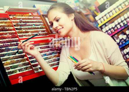 Smiling brunette girl picking différents crayons dans département artistique Banque D'Images