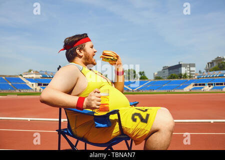 Gros homme mangeant un hamburger et de la bière tout en restant assis dans la formation au stade. L'alimentation, l'obésité. Banque D'Images