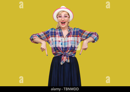 Portrait de femme mature élégant moderne surpris dans un style décontracté avec chapeau et lunettes debout, étonné à la caméra à l'intérieur et vers le bas. Banque D'Images