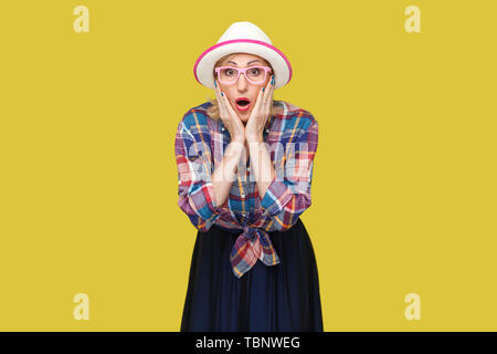 Portrait of young woman in casual élégant style avec chapeau et lunettes debout, toucher son visage et looking at camera with étonné face. je Banque D'Images