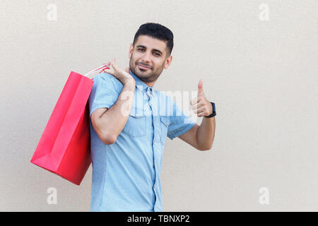 Les meilleures boutiques. Portrait de jeune homme barbu en chemise bleue avec sac shopping rouge permanent, looking at camera, sourire et Thumbs up. Piscine st Banque D'Images