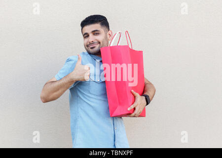 Cadeau étonnant. Portrait de jeune homme barbu satisfait en chemise bleu, rouge permanent holding Shopping bag, Thumbs up, looking at camera avec sourire. studi Banque D'Images