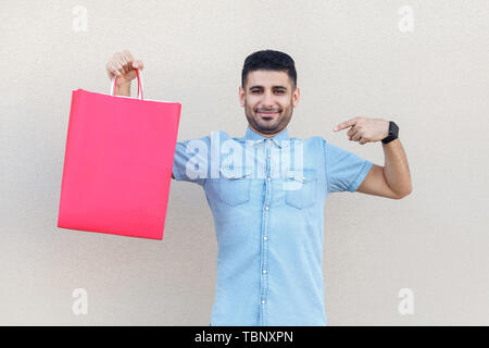 Portrait de beau jeune homme barbu en chemise bleue, tenant debout et pointant au red panier et looking at camera avec sourire. Piscine studi Banque D'Images