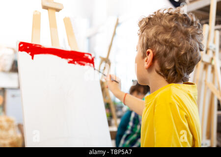 Avec la coloration rouge. Garçon aux cheveux blonds bouclés papier coloration rouge avec tout en maintenant la brosse de peinture Banque D'Images