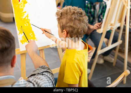 La coloration du papier. Joli garçon aux cheveux blonds bouclés holding pinceau peinture papier près de coloriage enseignant Banque D'Images