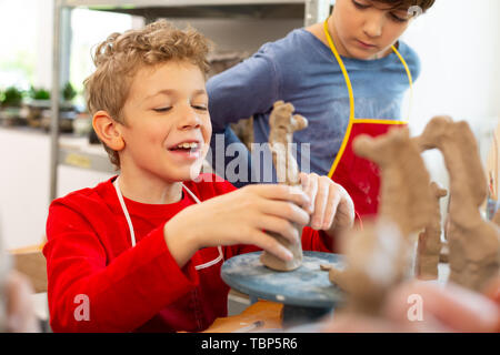 Garçon frisé de rire. Garçon aux cheveux blonds et bouclés tout en riant à modeler animaux au cours d'art Banque D'Images