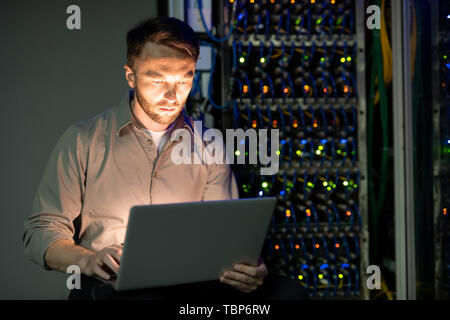 Les jeunes concentrés dans Server manager barbu shirt travail avec ordinateur portable dans une pièce sombre du centre de données Banque D'Images