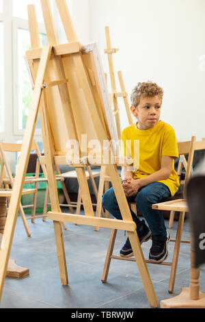 Cute funny boy. Cute funny boy wearing yellow t-shirt et jeans assis dans l'école d'art Banque D'Images