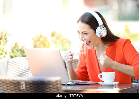 Excited woman wearing headphones regarder et écouter sur support ordinateur portable dans un café Banque D'Images