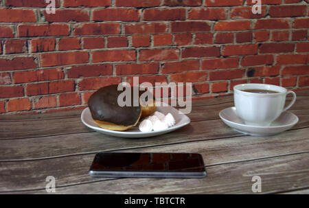Une plaque avec les beignets et les guimauves, une tasse de thé et un smartphone s'allonger sur une table de bois contre un mur de brique rouge. Banque D'Images