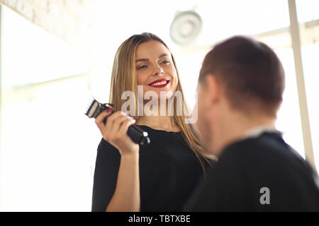 Smiling coiffure à l'aide de rasoir électrique à Client. Professional Hairstylist Holding Sharp Rasoir dans la main. Jeune homme rasage Barbier dans ce type. Femal Banque D'Images