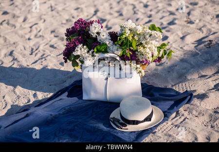 Bouquet de lilas sur la plage summes. Femme chapeau de paille, lunettes de soleil et sac élégant blanc sur une couverture bleue. Plage de sable fin. Banque D'Images