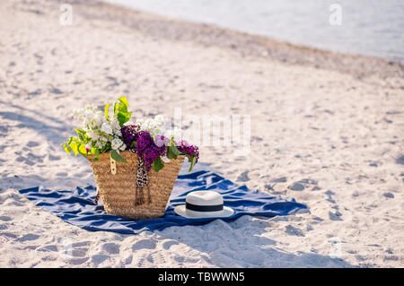 Bouquet de lilas dans un panier sur la plage summes. Femme chapeau de paille, lunettes de soleil et sac élégant blanc sur une couverture bleue. Plage de sable fin. Banque D'Images