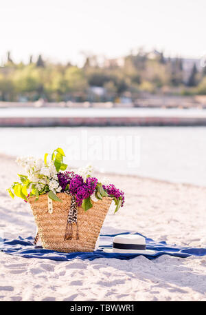 Bouquet de lilas dans un panier sur la plage summes. Femme chapeau de paille, lunettes de soleil et sac élégant blanc sur une couverture bleue. Plage de sable fin. Banque D'Images