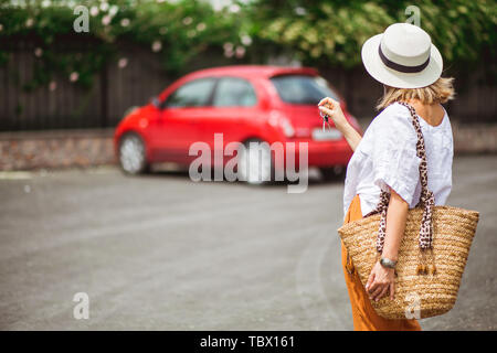 Femme en blanc se ferme et hat va à la petite voiture rouge. Louer ou acheter de l'auto concept. L'espace pour le texte. Voyager en voiture. La conduite. Achat nouvelle voiture. Banque D'Images