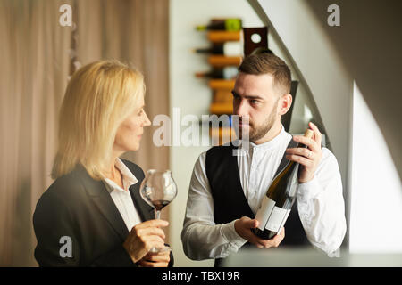 Portrait de sommelier aux teintes chaleureuses montrant la bouteille de vin à la femme au restaurant, copy space Banque D'Images