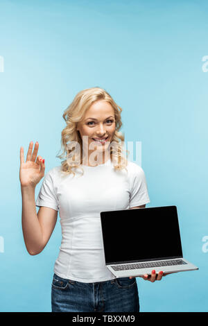 Happy blonde woman holding laptop avec écran blanc et montrant ok sign on blue Banque D'Images