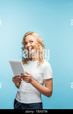 Happy blonde woman listening music des écouteurs pendant que holding digital tablet sur bleu Banque D'Images