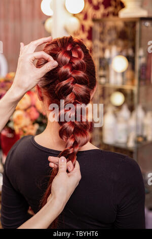 Belle, jeune fille rousse aux cheveux longs, coiffure tisse une tresse française, dans un salon de beauté. Soins des cheveux professionnels et la création de coiffures. Banque D'Images