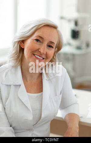 S'appuyant sur la table. Dame blonde agréable étant médecin expérimenté et le port de robe pendant son quart de représentant Banque D'Images