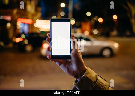 Woman's hand holding mobile phone avec affichage blanc sur fond de courts de nuit rue de la ville et de la route avec des voitures, close-up. Banque D'Images