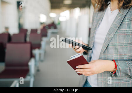 Jeune fille à l'aéroport, debout dans la zone d'attente avant le départ. Smartphone défilement holding, passeport et billet. Libre Banque D'Images