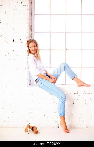 Coup de pleine longueur à maturité séduisante femme portant une chemise blanche et un jean tout en vous relaxant près de la fenêtre. Banque D'Images
