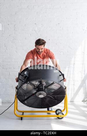 Bel homme excité debout près de mur blanc en face de ventilateur de soufflage Banque D'Images