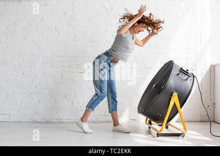 Excitée jeune femme en jean bleu debout avec les mains posées devant le ventilateur de soufflage Banque D'Images