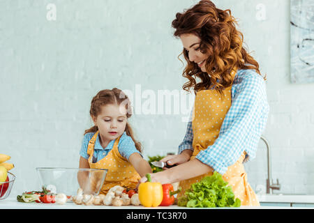 Smiling mother couper les légumes crus frais près de sa fille dans la cuisine Banque D'Images