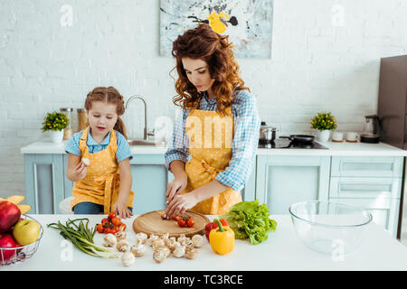 Mère en tablier à pois légumes crus coupe près de sa fille dans la cuisine Banque D'Images