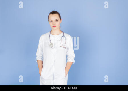 Jeune femme blonde médecin avec ses mains dans les poches avec stéthoscope sur son cou sur fond bleu avec l'espace pour le texte. Doctod examine l'appareil photo Banque D'Images