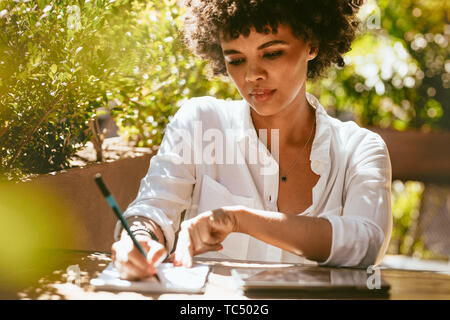 Freelancer faisant son travail à l'extérieur d'un café. L'écriture féminine africaine sur le bloc-notes assis à une table de café. Banque D'Images