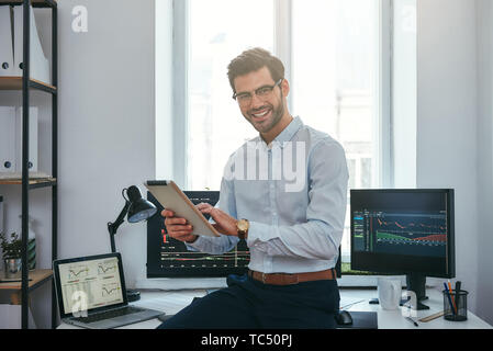 La réussite de l'entreprise. Cheerful young commerçant d'usure formelle est holding digital tablet and looking at camera avec sourire alors qu'il se tenait devant l'écran d'un ordinateur avec des graphiques dans l'office. Le marché de Forex. Concept commercial. Concept d'investissement Banque D'Images