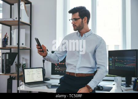 Stock Exchange. Jeune négociant ou un businessman concentré dans l'usure formelle et les lunettes est à la recherche dans les rapports sur son smartphone à l'objectif d'investissement tout en travaillant à son bureau. . Crypto-monnaies. Concept d'entreprise. Concept d'investissement Banque D'Images
