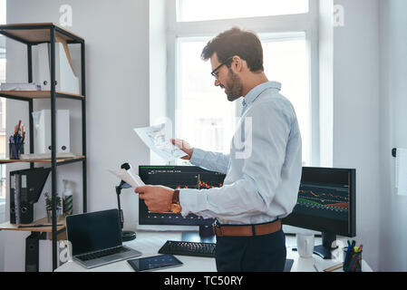 Bonne nouvelle heureux jeune homme d'affaires ou le commerçant à lunettes et à l'usure formelle à trading charts et souriant alors qu'il se tenait devant l'écran de l'ordinateur dans un bureau moderne. Courtier en valeurs. Le marché de Forex. Concept commercial Banque D'Images