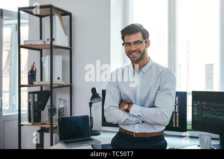 Heureux commerçant. Cheerful businessman dans les vêtements et les lunettes est en gardant les bras croisés, souriant et looking at camera while standing at office. Courtier en valeurs. Le marché de Forex. Concept commercial Banque D'Images