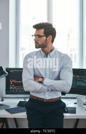 Jeune et intelligent. De photo verticale ou commerçant de vêtements formels et les lunettes à la route et à garder les bras croisés en se tenant debout à l'office. Courtier en valeurs. Le marché de Forex. Concept commercial Banque D'Images