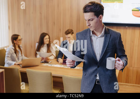 Un jeune homme élégant dans un veston et une chemise avec une tasse de café dans sa main se trouve et lit des documents sur le contexte de travail des collègues de l'off Banque D'Images