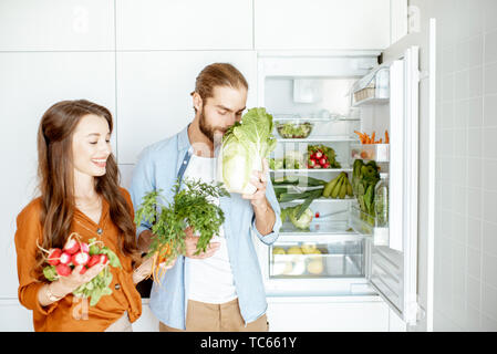 Portrait d'un jeune et heureux couple standing avec légumes frais près du réfrigérateur plein de produits sains à la maison Banque D'Images