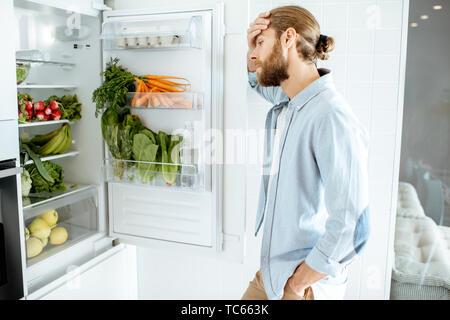 Homme triste de choisir des produits à cuire, debout près du réfrigérateur plein de légumes à la maison Banque D'Images