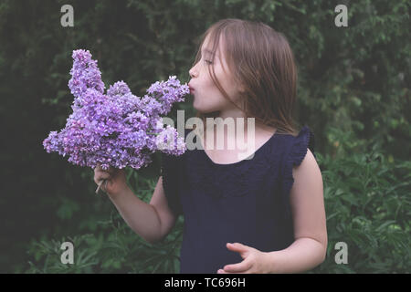 Cute little girl kissing lilas. Enfant s'amusant à l'extérieur. Petit enfant bénéficiant, aimant la nature. Belle botte, bouquet de fleurs violettes. Portrait de la personne, sciences naturelles, floral background Banque D'Images