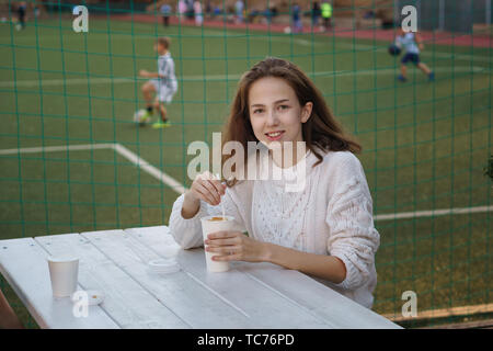 High school student boire du café sur la terrasse d'un café de l'école. Elle sourit et pose pour appareil photo. Terrain de football de l'arrière-plan. Temps pour Banque D'Images