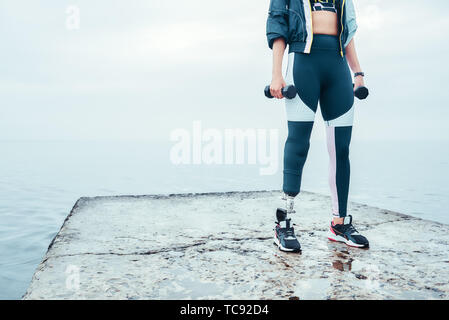 L'entraînement avec haltères. Portrait de femme handicapée dans les vêtements de sport avec haltères holding prothèse de jambe en position debout en face de la mer. Mobilité sportif. Mode de vie sain. Workout Banque D'Images